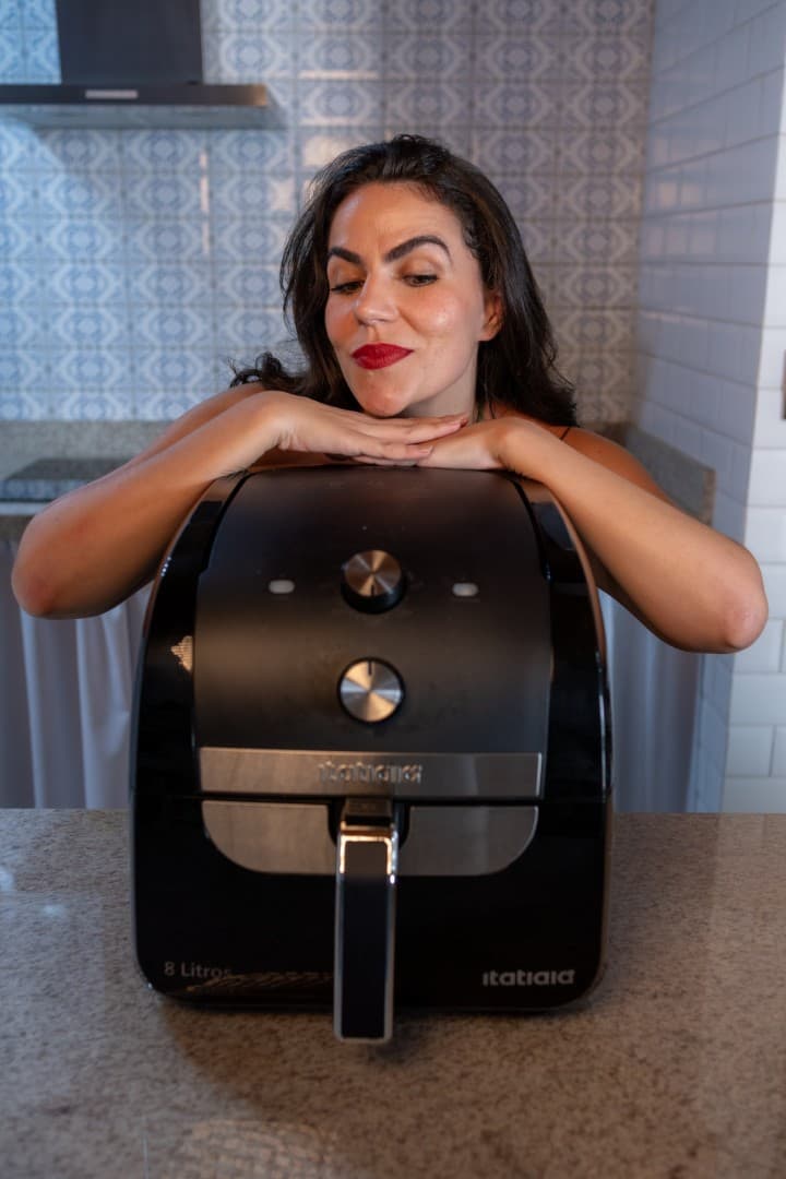 Cecília apoiada em uma air fryer preta na cozinha.