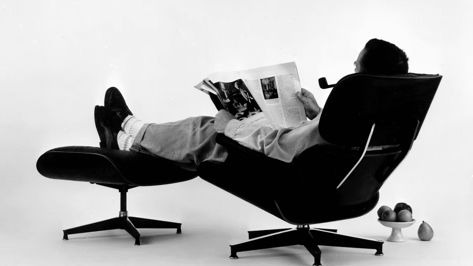 Pessoa lendo na Cadeira Eames Lounge com os pés na otomana