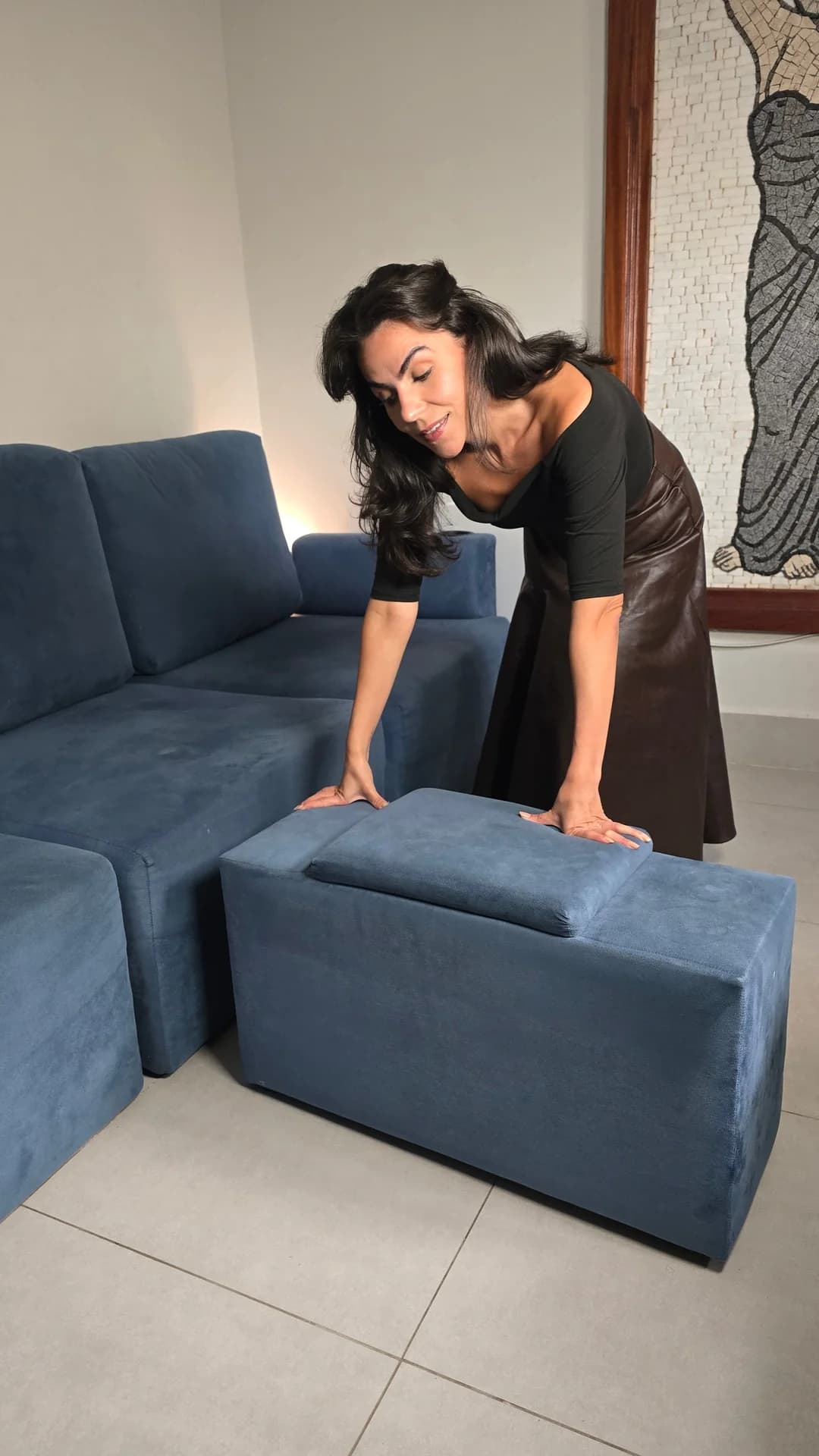 Cecília com as mãos apoiadas no puff do sofá modular DAMIE azul, na sala de casa